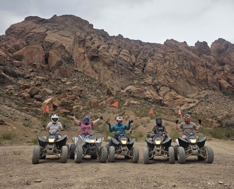 Riders in helmets on ATVs celebrate in a Nevada desert near Las Vegas with rocky hills, orange flags, and ATV tour vibes.