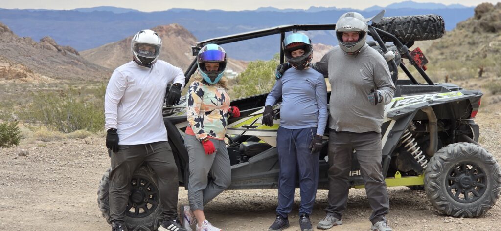 Group with helmets in casual gear stands by RZR off-road vehicle in Nevada desert near Las Vegas on ATV tour, mountains behind.