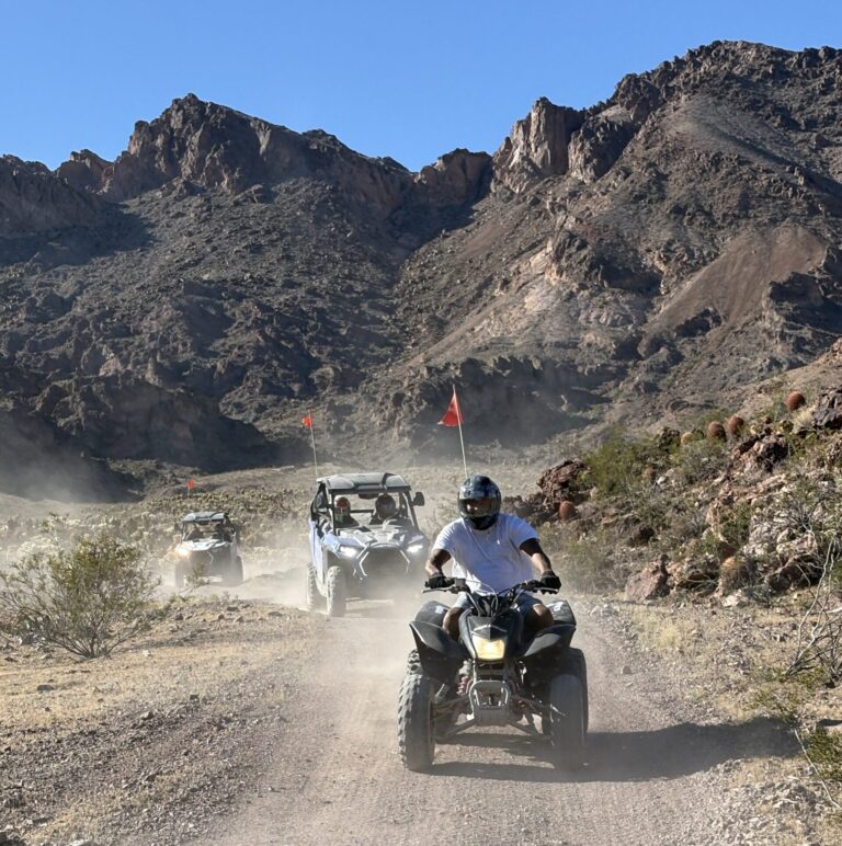 Helmeted rider leads ATV tour on a Nevada desert trail near Las Vegas, with RZR vehicles kicking up dust in rocky mountains.
