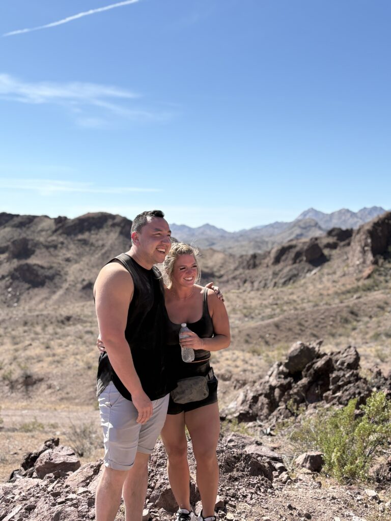 Smiling couple in activewear on a rocky Nevada trail near Las Vegas with mountains; woman holds water bottle.