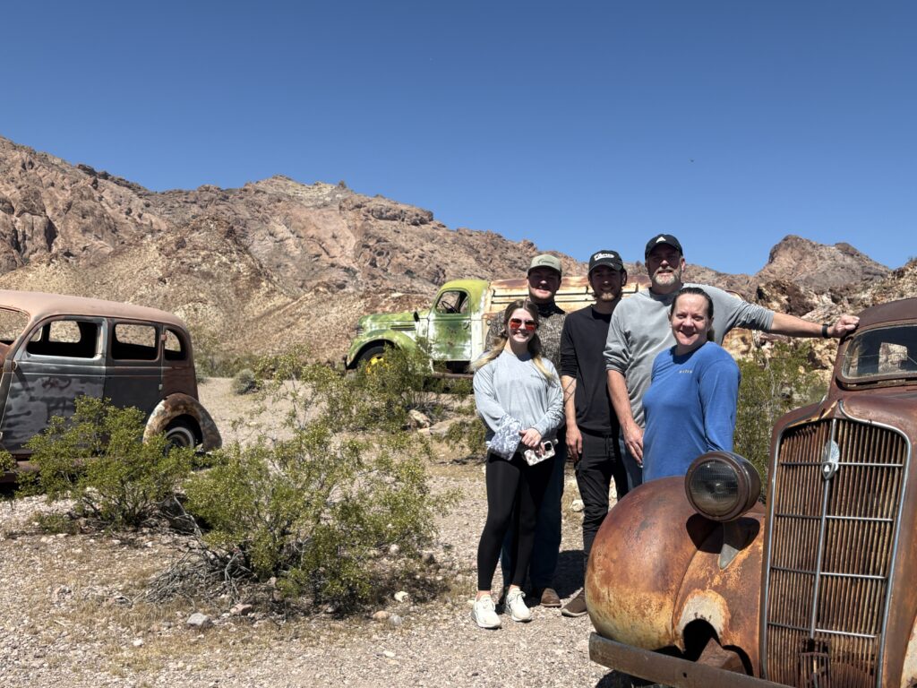 Group smiling by rusty vintage cars in Nevada desert near Las Vegas, perfect for Ghost Town sightseeing or ATV tours.