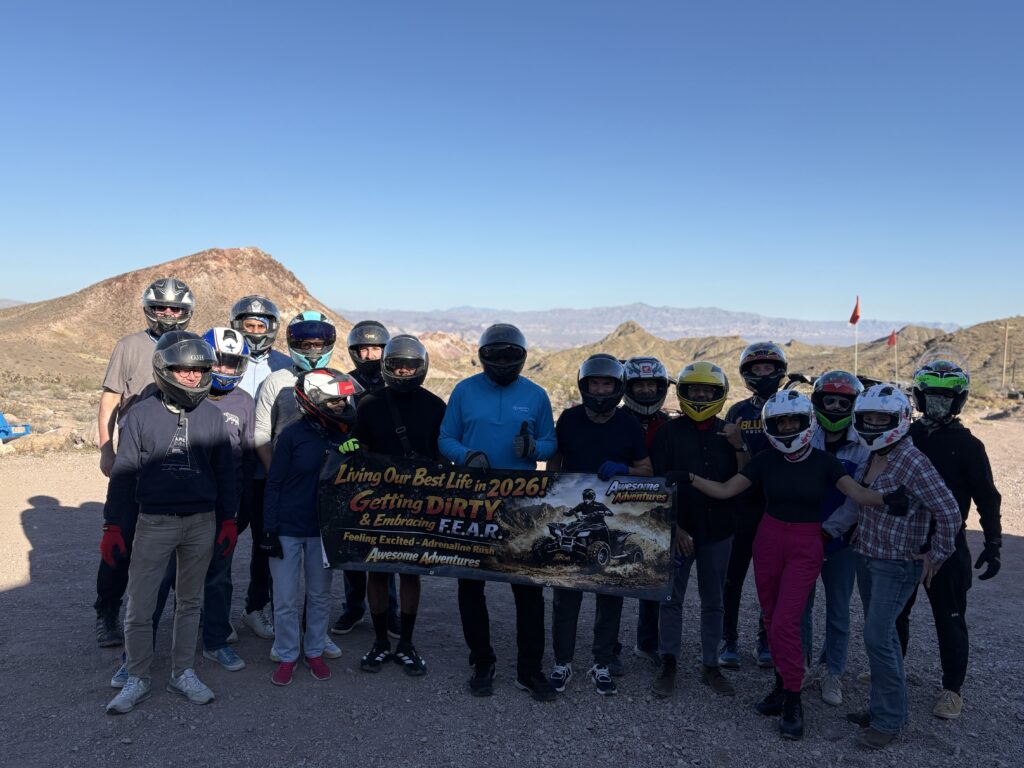 Group in helmets near Las Vegas on rocky Nevada terrain, holding 2026 banner after an ATV tour with hills and blue sky behind.