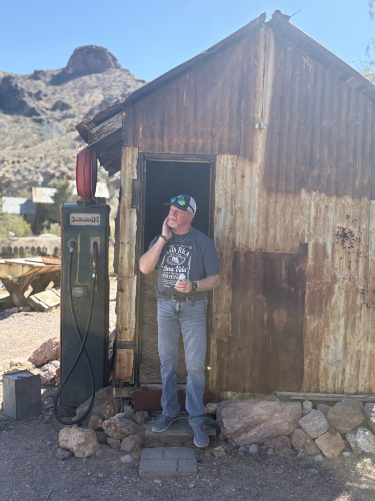 Man stands by an old gas pump at a weathered Nevada ghost town shed, mountains behind—perfect for Las Vegas ATV tours.