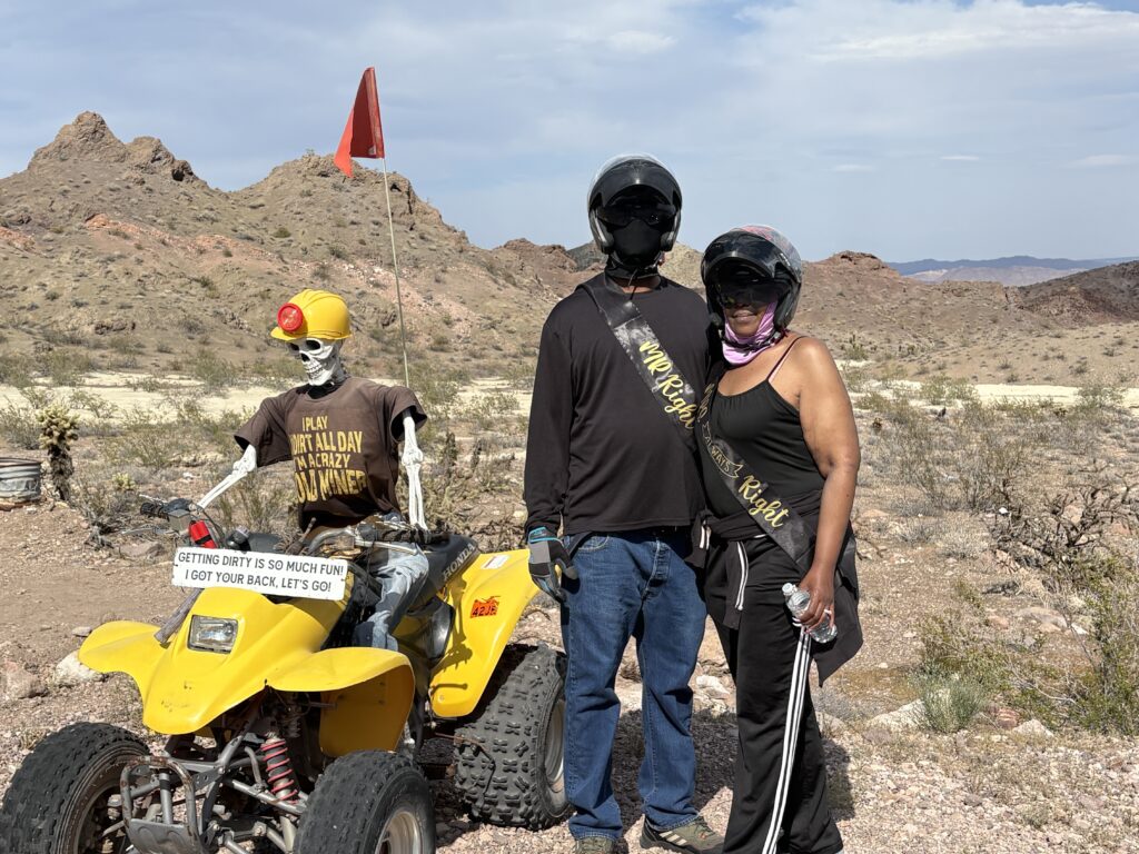 Two in helmets pose near a skeleton on ATV in Nevada desert; Ghost Town sightseeing & ATV tours near Las Vegas.