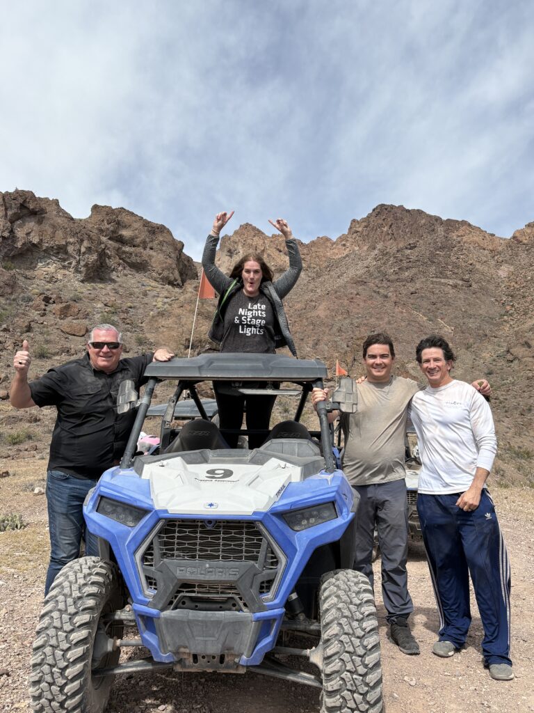 Adventurers pose by a blue RZR on a rocky Nevada trail near Las Vegas, with mountains behind, enjoying ATV off-road fun.