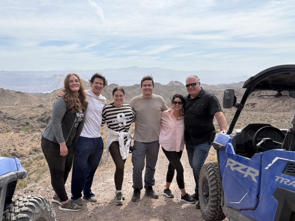 Smiling group on a rocky Nevada trail with two ATVs, desert hills, and mountains—Las Vegas RZR off-road adventure.