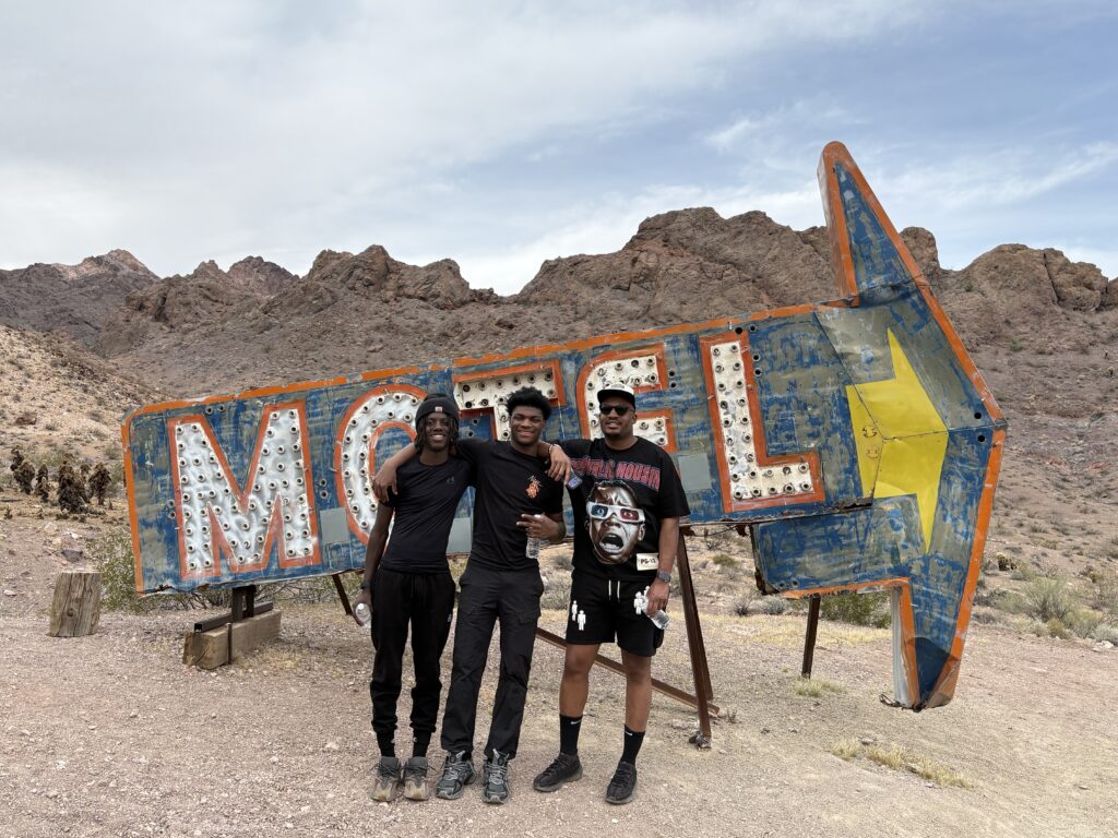 Three men pose by a weathered MOTEL sign in the Nevada desert near the Colorado River, perfect for ghost town sightseeing.