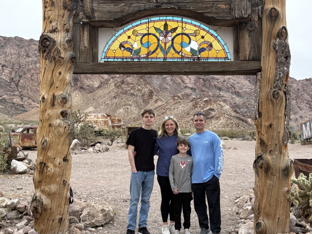 Smiling family under rustic arch with stained glass in Nevada desert, near Las Vegas Ghost Town and old vehicles, perfect for sightseeing.