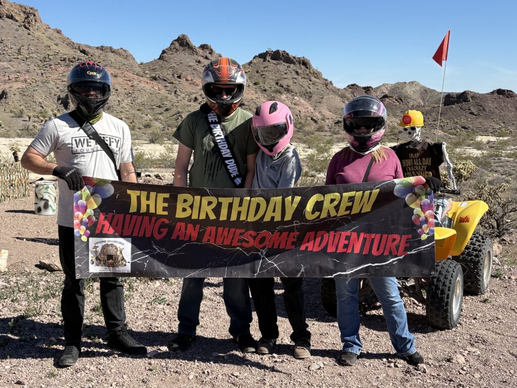 Four adventurers with helmets hold a “Birthday Crew” banner near an ATV in the Nevada desert on a Las Vegas off-road tour.