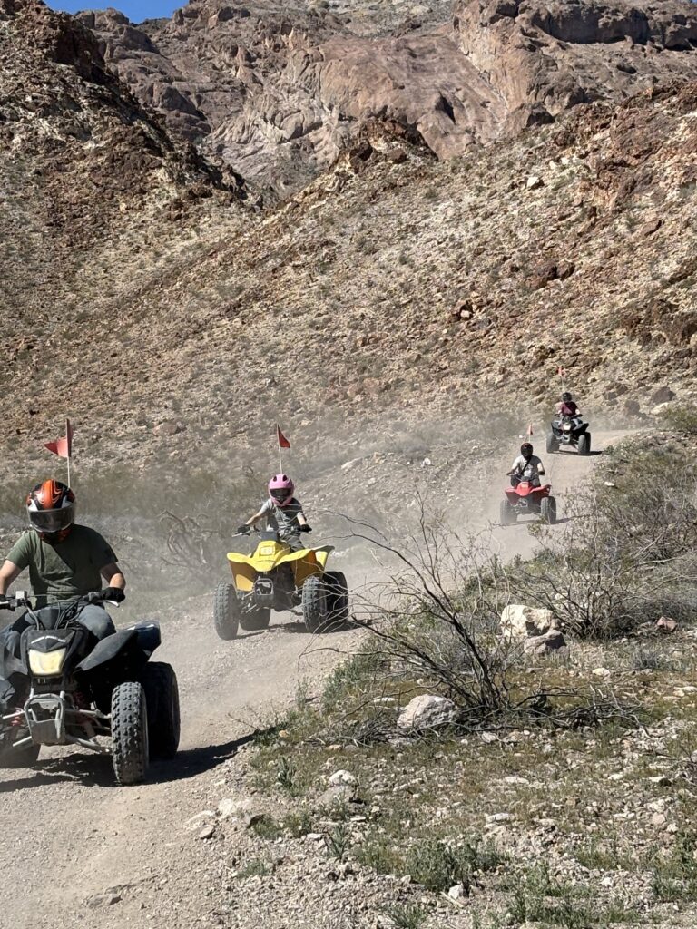 ATV tour near Las Vegas: four riders in helmets explore a dusty Nevada desert trail, mountains and desert scenery behind.