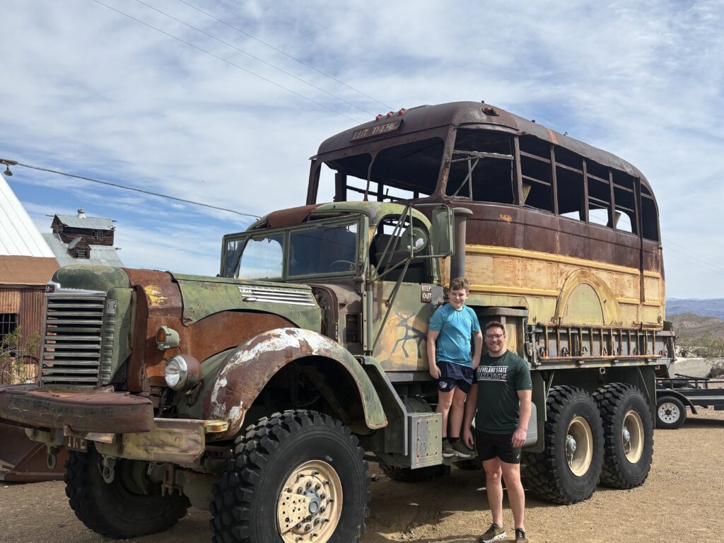 Smiling duo by a rusty vintage military truck with bus frame in the Nevada desert—perfect for Las Vegas Ghost Town sightseeing.