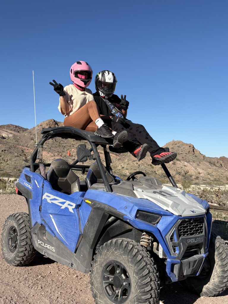 Adventurers in helmets pose on a Polaris RZR during an ATV tour near Las Vegas, with Nevada hills and blue skies behind them.