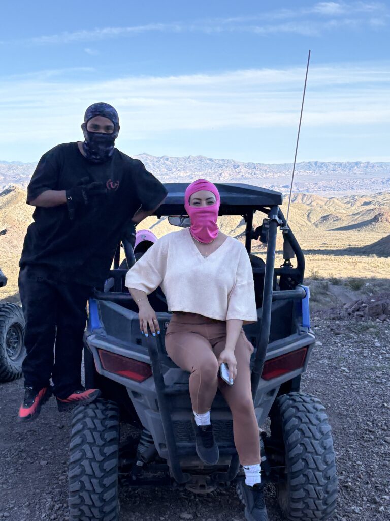 Adventurers in face coverings enjoy a Nevada ATV tour near Las Vegas, posing with an RZR on a rocky desert trail under blue skies.
