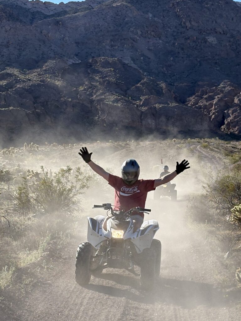 ATV riders explore a dusty Nevada desert trail near Las Vegas, raising hands with rocky hills behind during an off-road tour adventure.