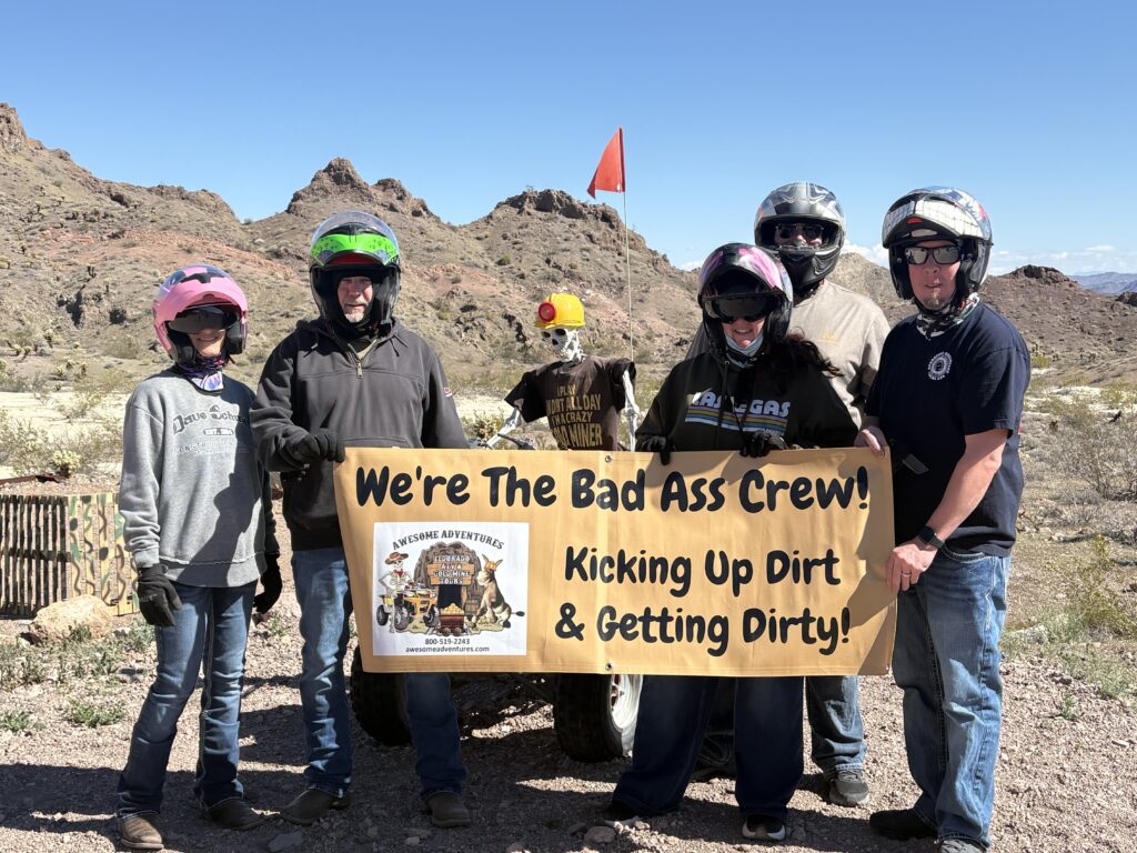Five riders in helmets pose by a "Bad Ass Crew" banner during an ATV tour in the Nevada desert near Las Vegas and the Colorado River.