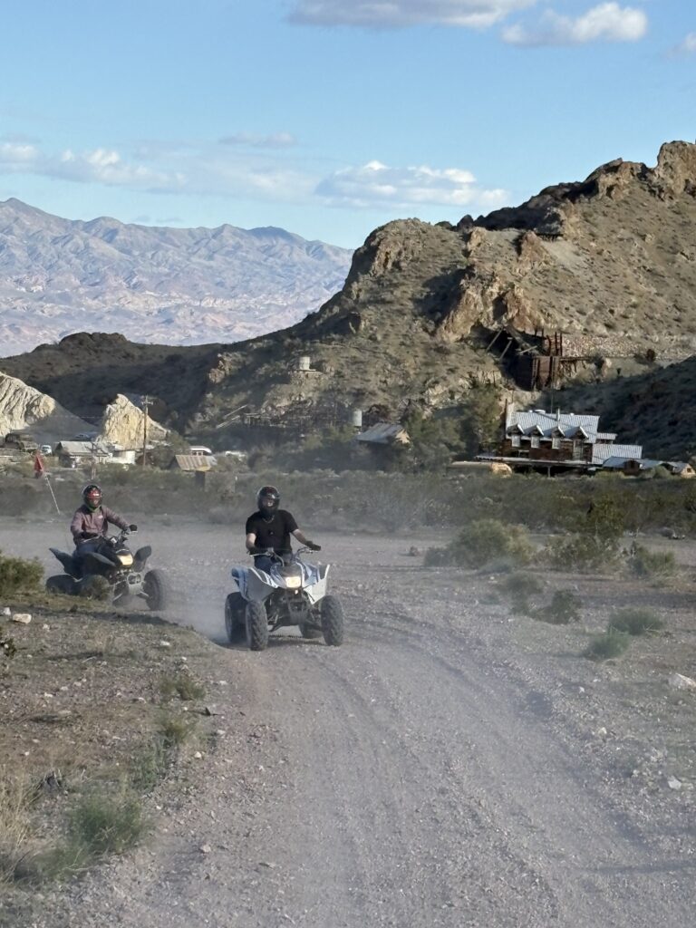 ATV riders explore a dusty Nevada desert trail near Las Vegas, passing ghost town buildings and rocky hills under partly cloudy skies.