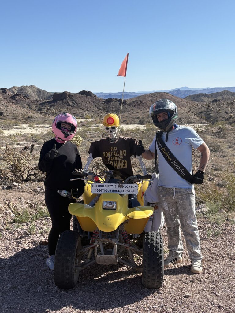 Three friends in helmets pose by a yellow ATV near Las Vegas; rocky Nevada desert and blue sky hint at an epic ATV tour adventure.
