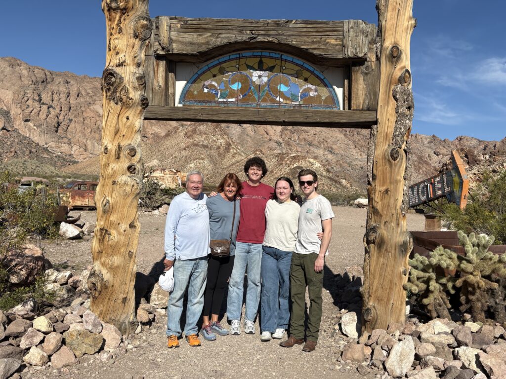 Group smiles under a rustic arch in a Nevada desert ghost town near Las Vegas, with old vehicles and rocky hills in the background.