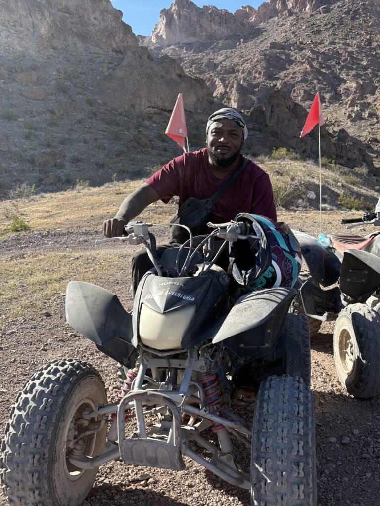 Man in red shirt rides black ATV on rocky Nevada trail, mountains and red flags behind, perfect for Las Vegas ATV tours.
