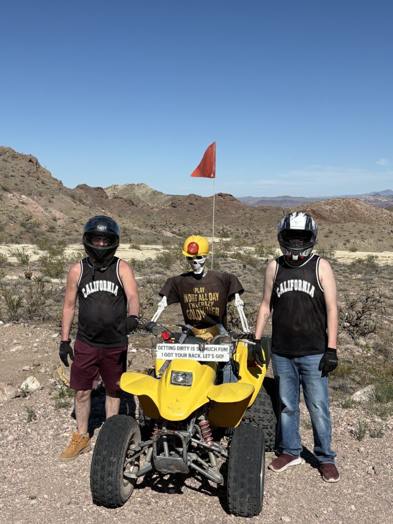 Adventurers in black tank tops by a yellow ATV, skeleton rider, Nevada desert backdrop—Las Vegas ATV tour near Colorado River.