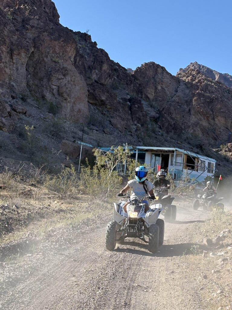 ATV riders on a dusty Nevada trail near rocky hills and a ghost town house, perfect for Las Vegas RZR off-road tours.