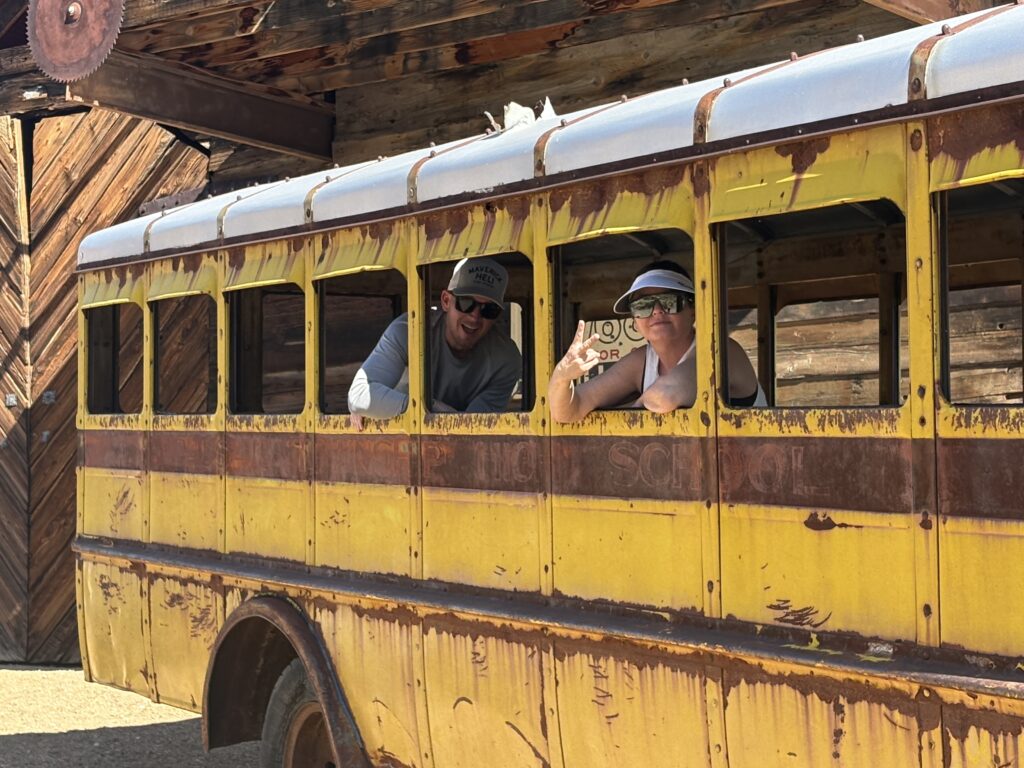 Two people wave from a rusty yellow school bus in sunny Nevada near a wooden building—perfect for Ghost Town sightseeing.