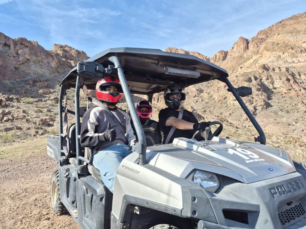 RZR off-road ride near Las Vegas: three people in helmets sit in a gray ATV on rocky Nevada terrain under blue sky.