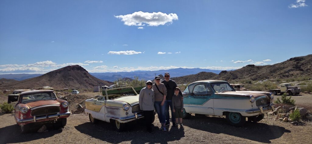 Smiling group by vintage cars in Nevada desert near rocky hills and blue sky—perfect for Las Vegas ghost town sightseeing.