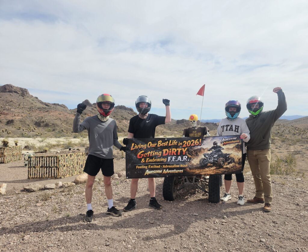 Four friends in helmets pose with a banner by an ATV near Las Vegas, Nevada, after a RZR off-road ride in the desert.