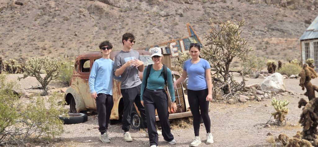 Group by a rusty car and motel sign near Nevada desert, enjoying sunny Ghost Town sightseeing with cacti, rocky hills, and sunglasses.