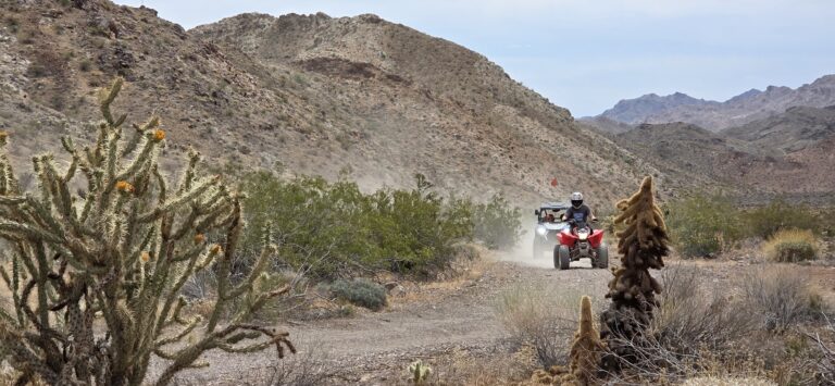 ATV riders explore a dusty desert trail near Las Vegas, Nevada, passing rocky hills, cacti, and shrubs under cloudy skies.