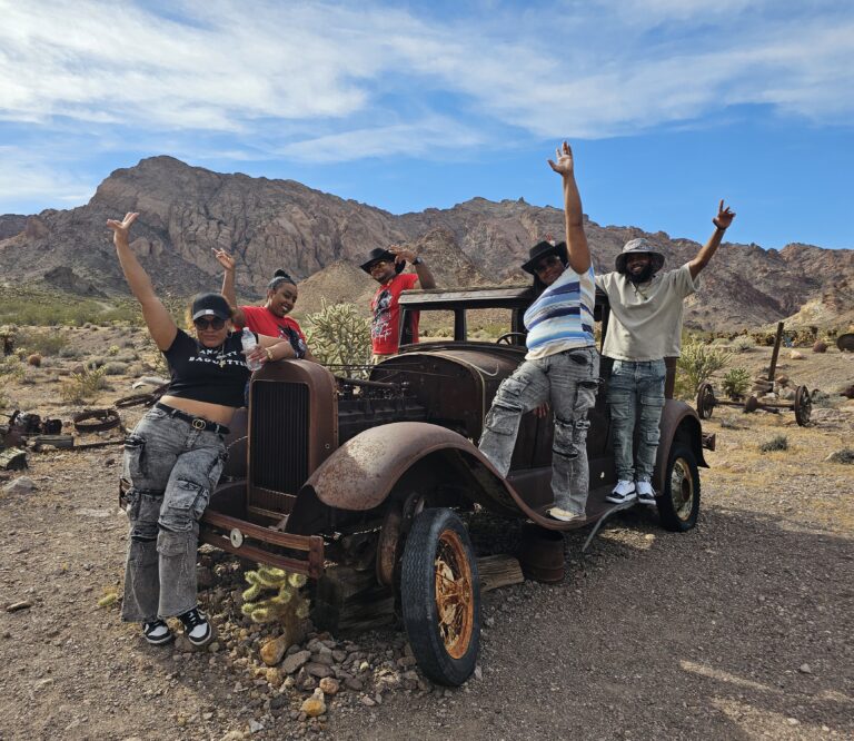 Friends celebrate by a rusty vintage car in Nevada’s desert, with Las Vegas mountains—perfect for Ghost Town sightseeing.