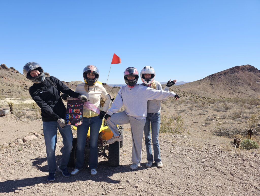 Four friends on a Las Vegas ATV tour pose by a yellow quad in the Nevada desert, holding a birthday sign; red flag waves.