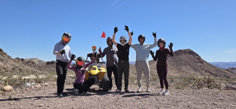 Group wearing helmets and gloves celebrates by a yellow ATV in Nevada near Las Vegas on an off-road tour; desert mountains behind.