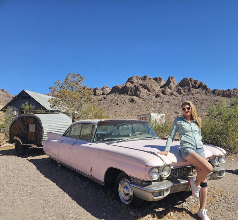 Woman in sunglasses leans on vintage pink car near Nevada ghost town, mountains, and trailer, perfect for ATV or RZR tours.