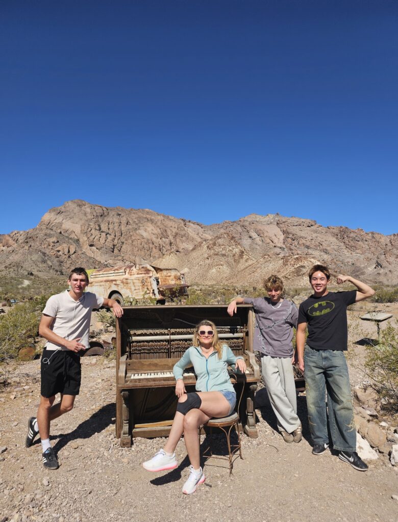 Four friends pose by a broken piano in the Nevada desert near Las Vegas, rocky mountains behind, perfect for ghost town sightseeing.