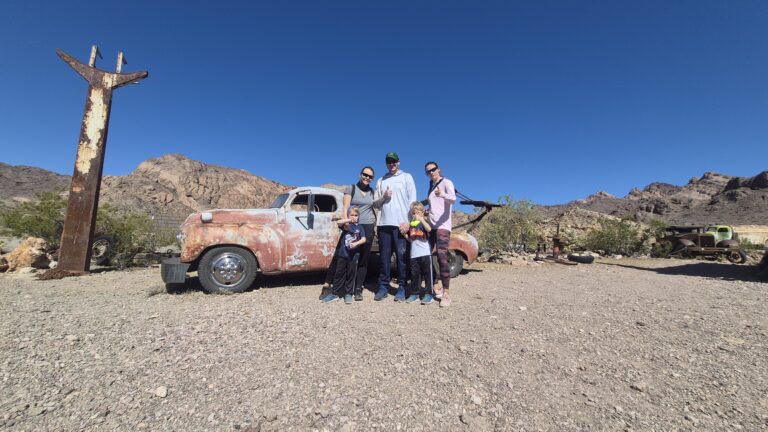 Family of five poses by a rusty car near Las Vegas, Nevada desert mountains—perfect for Ghost Town sightseeing adventures.