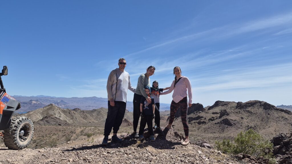 Four people smile atop a rocky Nevada hill with mountain views, clear sky, and an ATV during an off-road tour near Las Vegas.