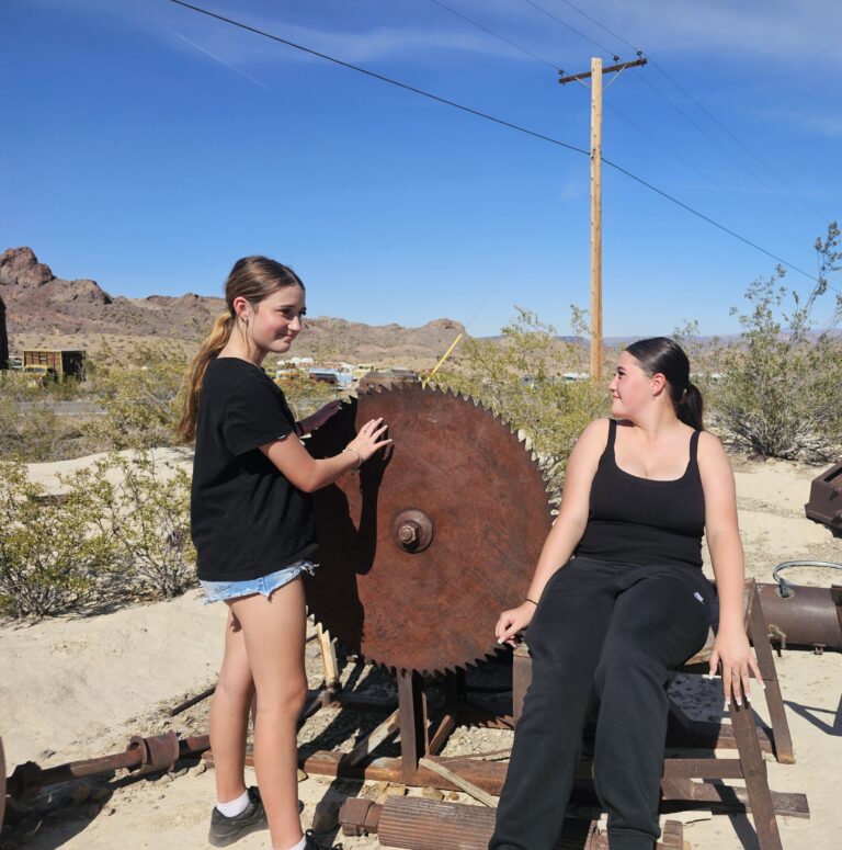 Two women in black outfits pose by a rusty saw blade in a Nevada desert near Las Vegas mountains, ideal for ghost town sightseeing.