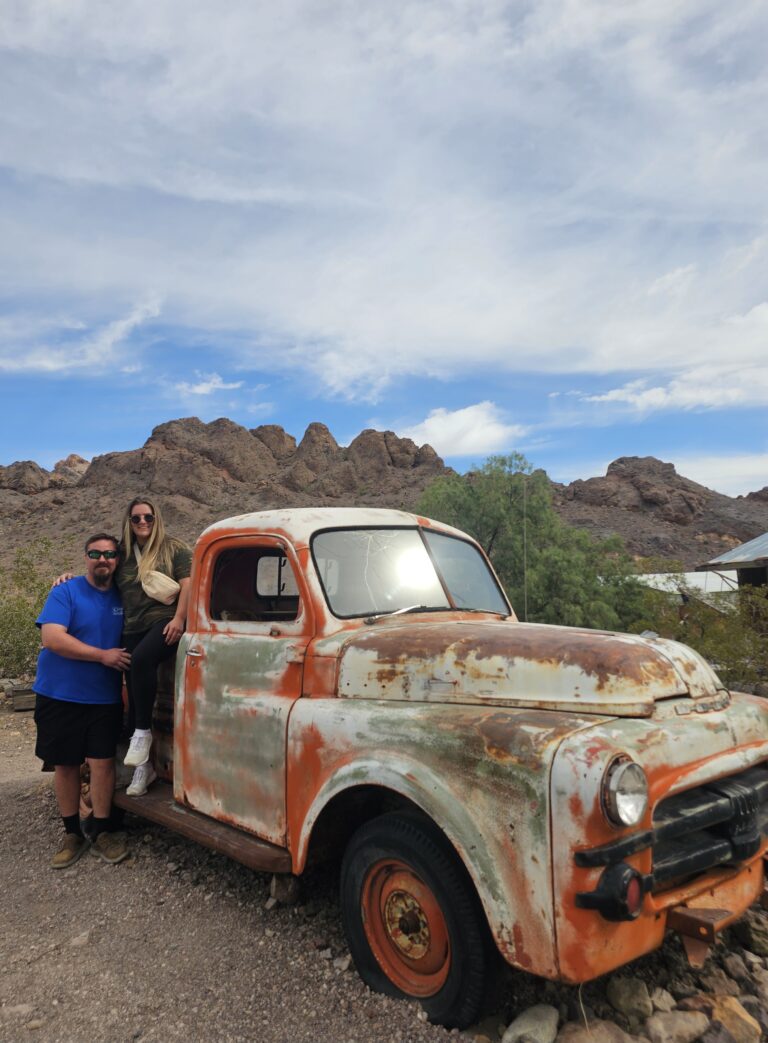 Smiling couple poses by a rusty pickup in Nevada desert near Las Vegas, perfect for Ghost Town sightseeing adventures.