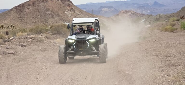 Nevada ATV tour: Two helmeted riders in a black RZR blaze a dusty trail through rocky desert hills near Las Vegas.