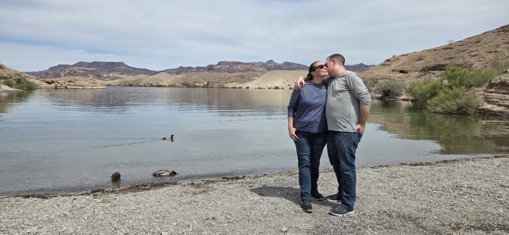 Couple embraces on Nevada’s Colorado River shore, smiling as ducks swim by; scenic hills and cloudy sky reflected in calm water.