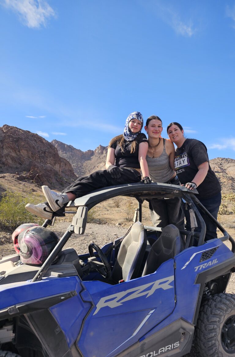 Three women enjoy an ATV RZR ride near Las Vegas in a Nevada desert, posing by rocky hills under clear sunny skies.