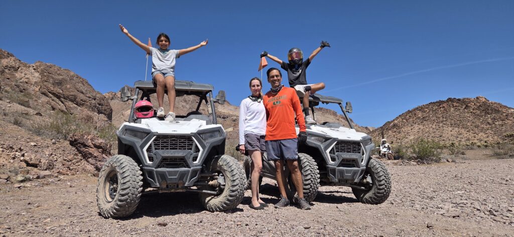 ATV tour near Las Vegas: two RZRs on rocky terrain, family poses with arms raised, Nevada hills and blue sky in the background.