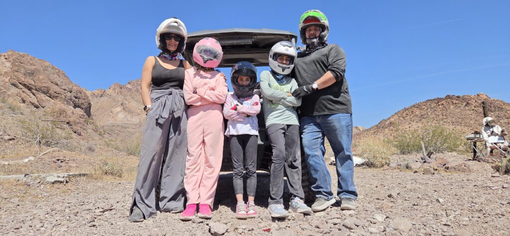 Five people in helmets and gear pose by an ATV in Nevada desert near Las Vegas, ready for RZR off-road tours or Ghost Town sightseeing.