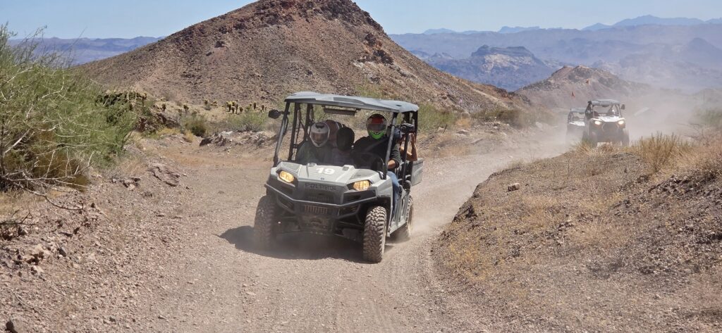 Adventurers ride RZR off-road vehicles near Las Vegas on a dusty Nevada desert trail with mountain and Colorado River views.