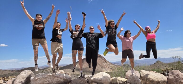 Seven women jump with joy on a rocky Nevada ledge near the Colorado River, enjoying Ghost Town sightseeing and scenic views.