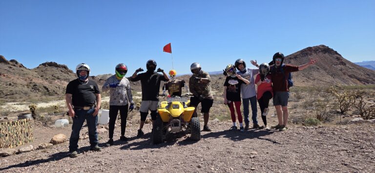 Seven riders in helmets pose by a yellow ATV with red flag during a sunny Las Vegas Nevada desert ATV tour near rocky hills.