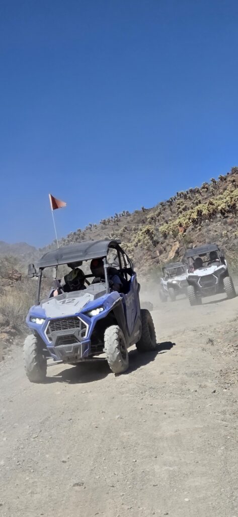ATV tour near Las Vegas: three off-road vehicles race on a dusty Nevada trail by rocky desert and blue sky, kicking up dust.