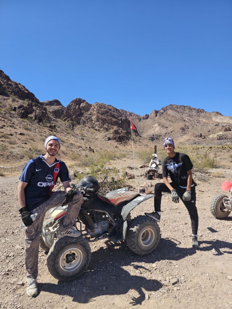 Adventurers with gloves and headbands pose by an ATV on a Nevada desert trail near Las Vegas, with RZR tours in the background.
